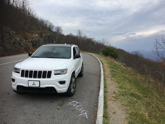 A white Jeep Grand Cherokee parked on the shoulder of a scenic mountain road during an overcast day.