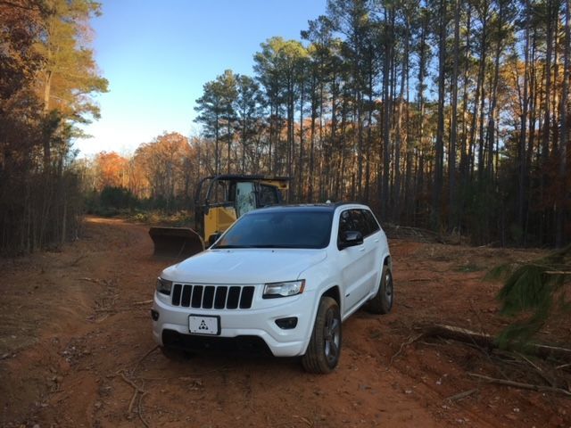 A white Jeep Grand Cherokee parked on a dirt path in a forest next to a piece of yellow construction equipment.