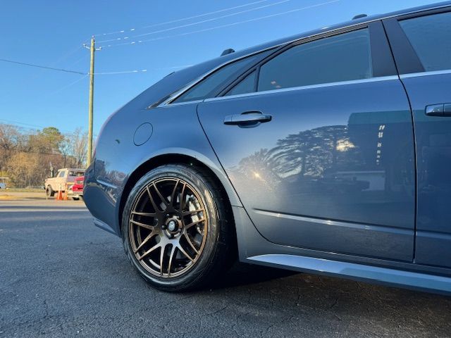 A low-angle view of the rear side and bronze alloy wheel of a blue station wagon parked on an asphalt lot.
