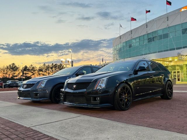 Two Cadillac CTS-V wagons parked on a brick plaza in front of a modern glass building at sunset.