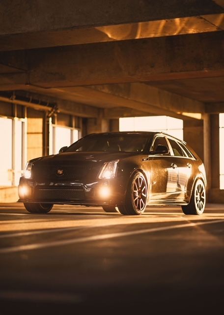 A dark-colored Cadillac station wagon parked in a dimly lit concrete parking garage, illuminated by golden sunlight.