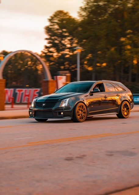 A dark gray Cadillac CTS-V wagon with bronze wheels driving on a paved road during sunset near a brick arched entrance.