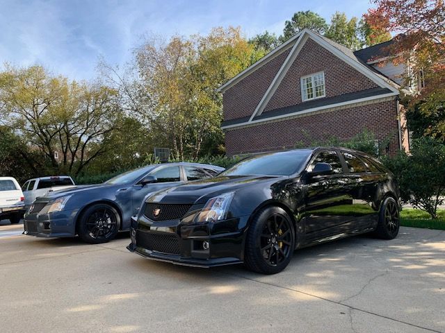 Two Cadillac CTS-V wagons, one black and one gray, parked on a driveway in front of a brick house on a sunny day.