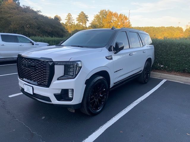 White GMC Yukon SUV parked in a paved parking lot on a sunny day with green landscaping in the background.