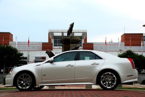 A white Cadillac parked in front of a brick building with a large wolf statue in the background.