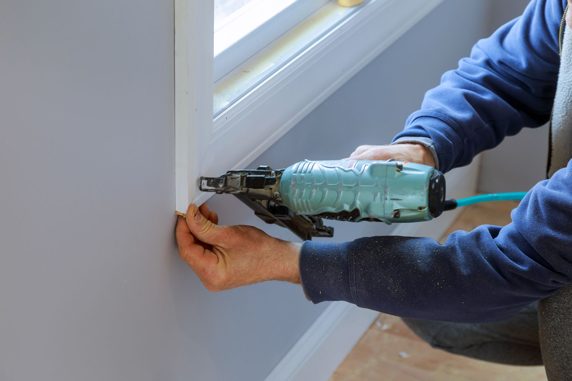 A person uses a teal pneumatic nail gun to attach white window casing to a wall.