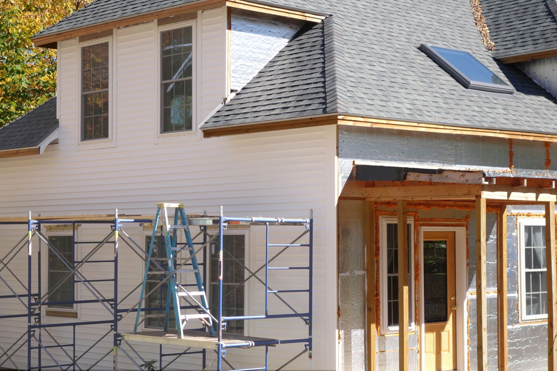 A house under construction with light-colored siding, scaffolding in front, a gray shingled roof, and a skylight.