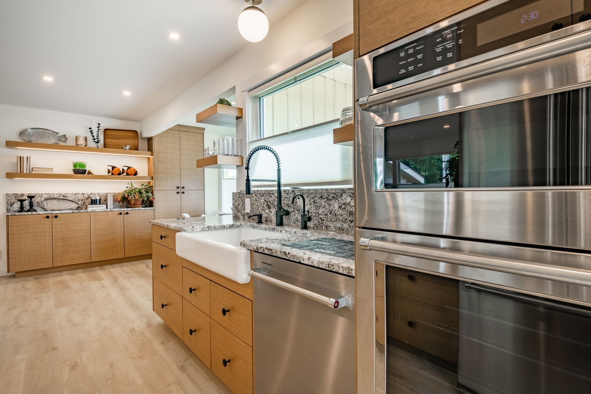 A modern kitchen featuring light wood cabinets, an apron-front sink, granite countertops, and stainless steel appliances.