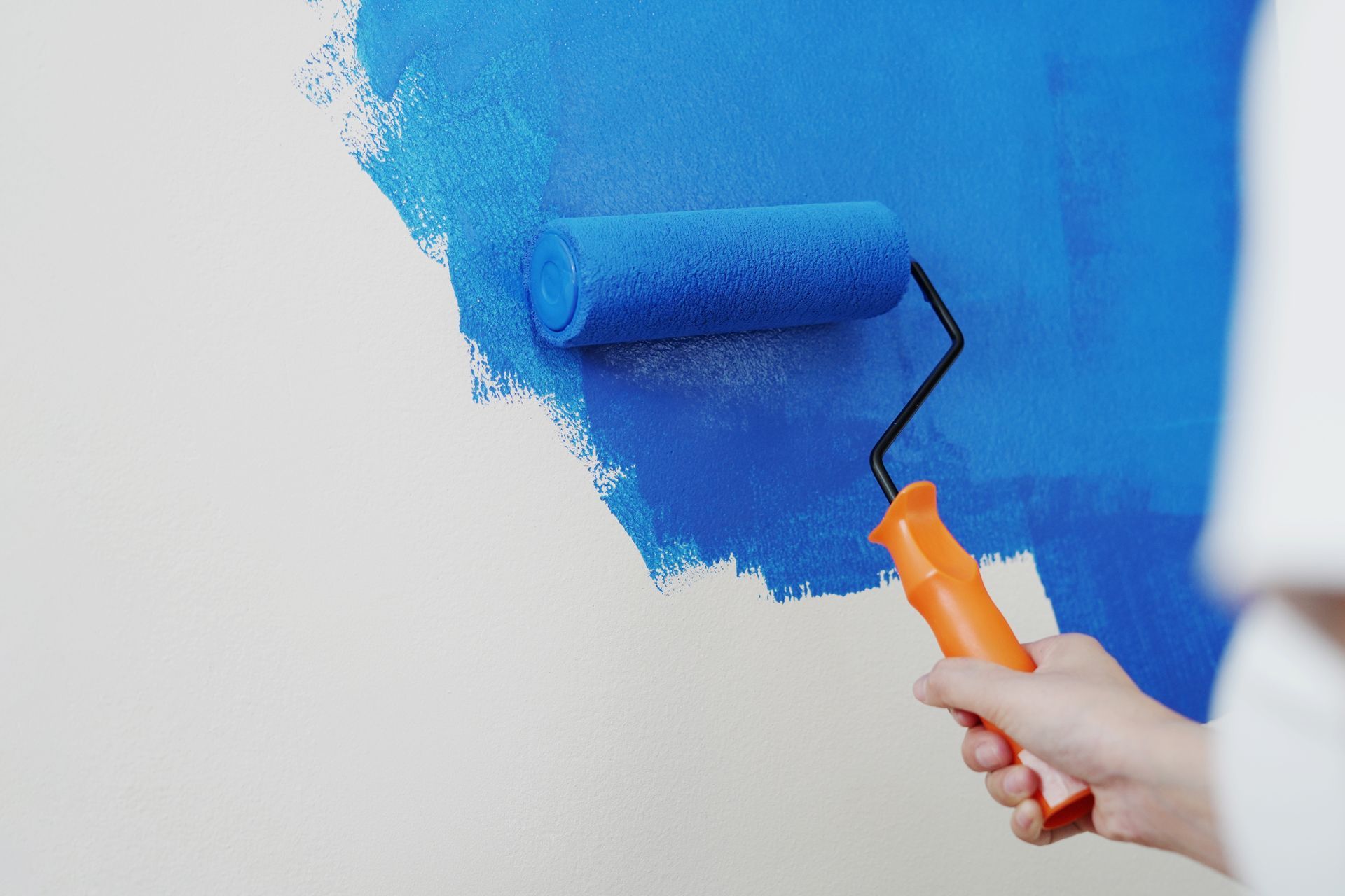 A hand holds an orange-handled paint roller, applying bright blue paint to a white wall.