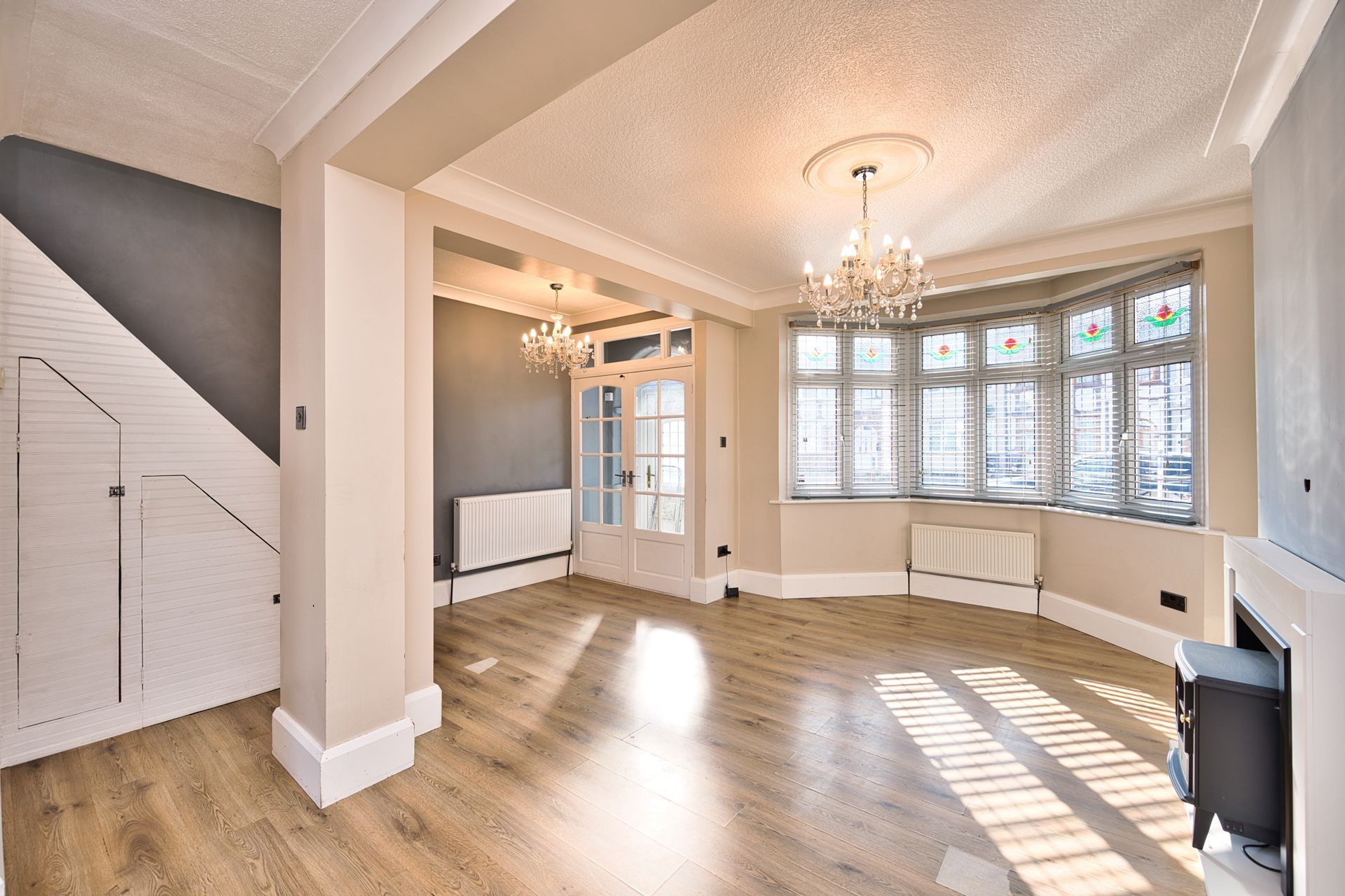 Bright open living room with light wood floors, gray accent walls, a large bay window, and ornate crystal chandeliers.