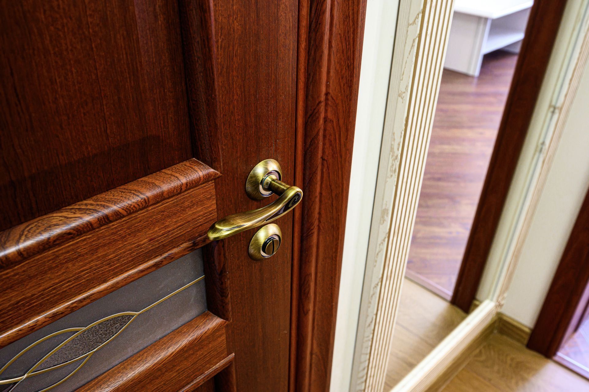 Close-up of a wood-textured door with an antique gold handle, partially open to reveal a room with hardwood flooring.