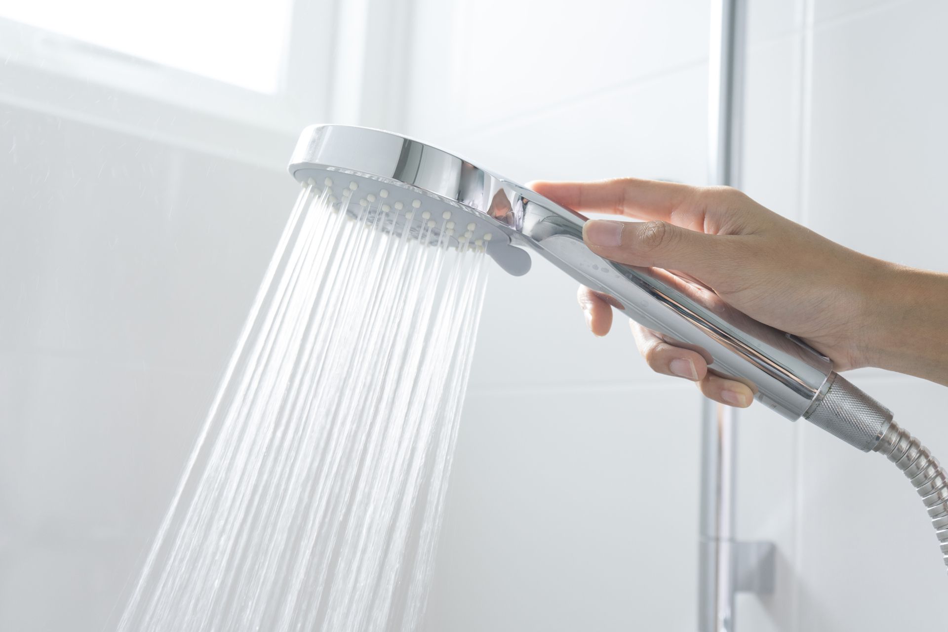 A hand holds a chrome handheld showerhead with water spraying out in a steady, focused stream against a tiled background.