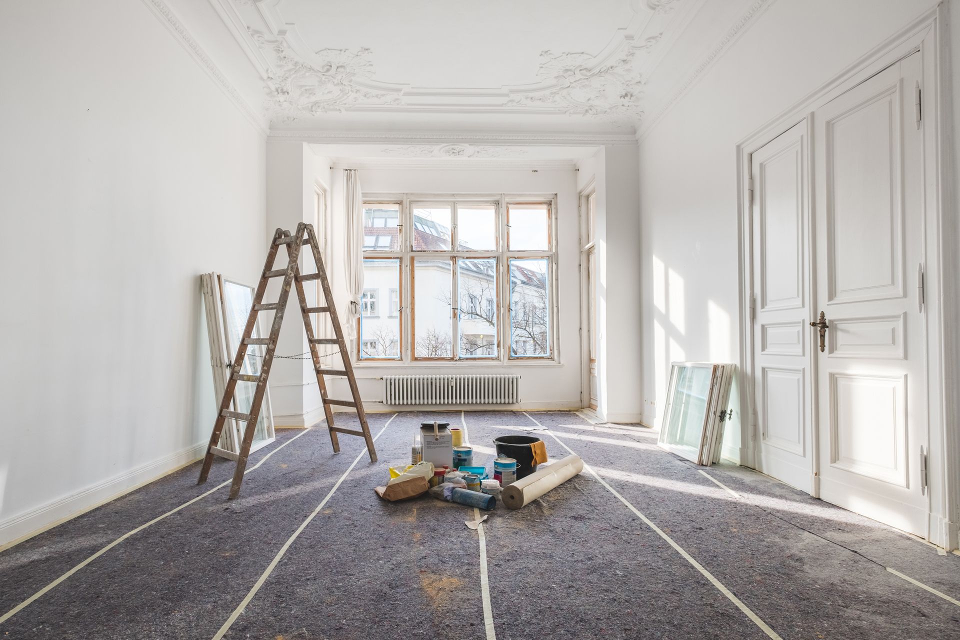 A bright room under renovation with a ladder, painting supplies, and protective floor covering.