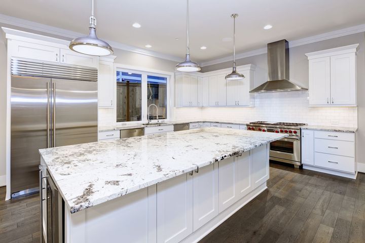 A modern kitchen with white cabinets, a large granite island, stainless steel appliances, and dark hardwood floors.