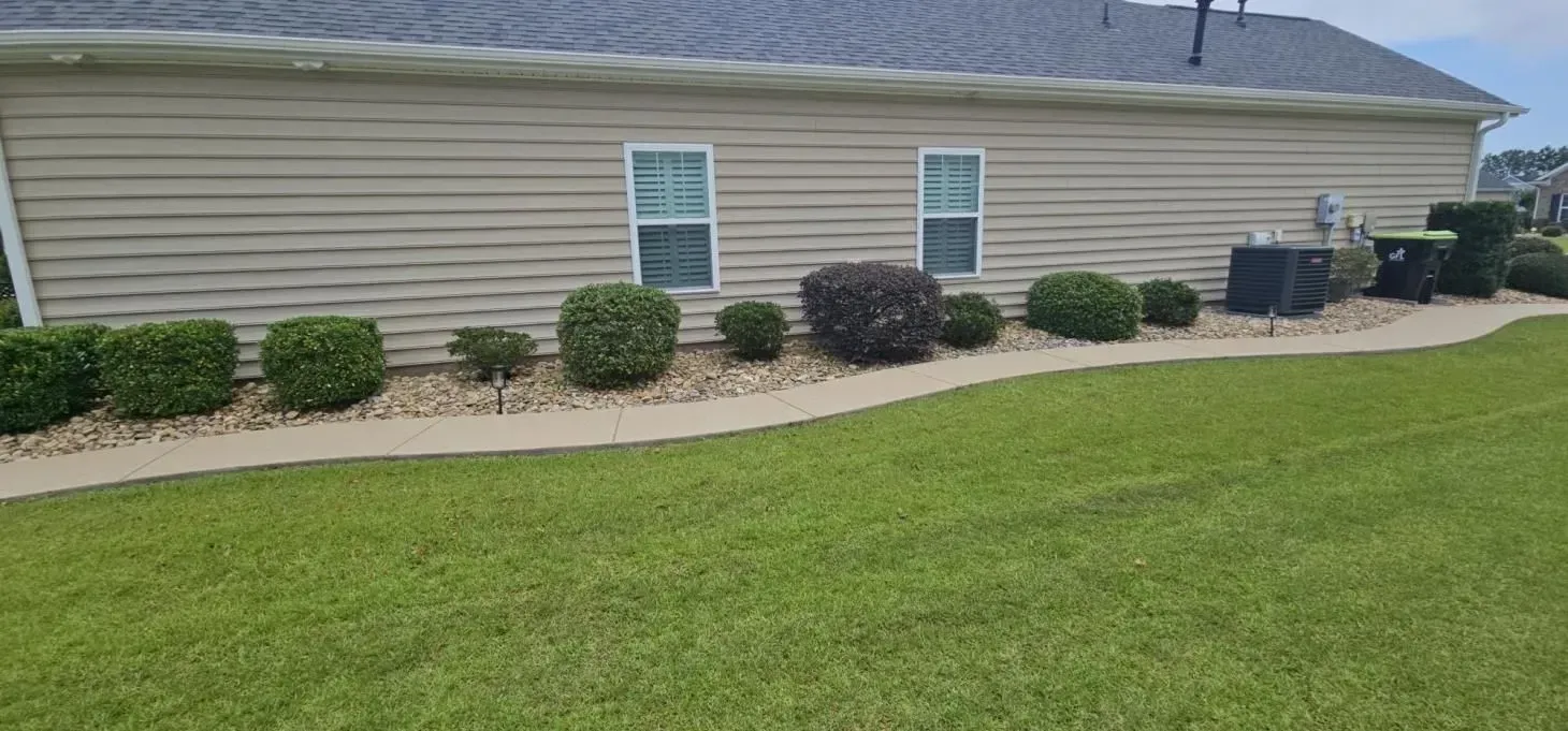 Side of a beige house with green bushes, grass, and a concrete walkway.