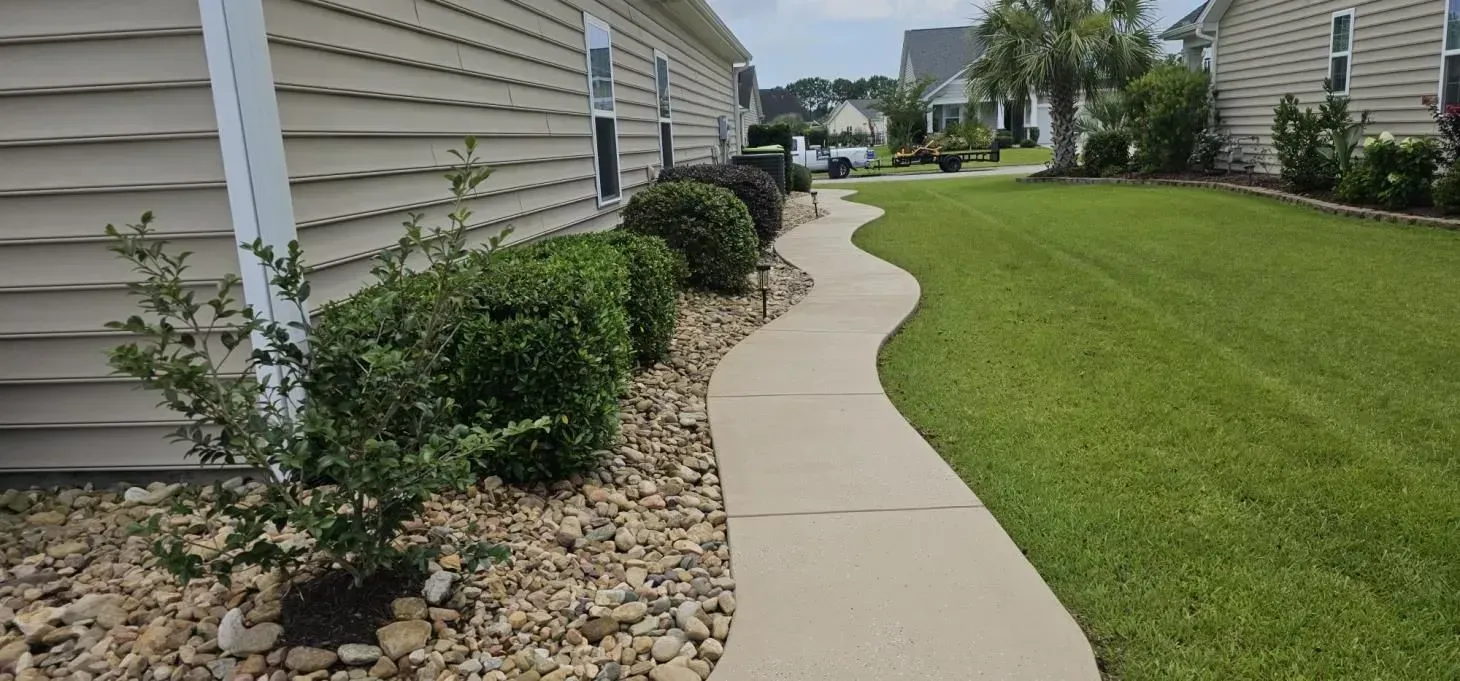 A winding concrete sidewalk leads through a green lawn and garden beds beside a house.