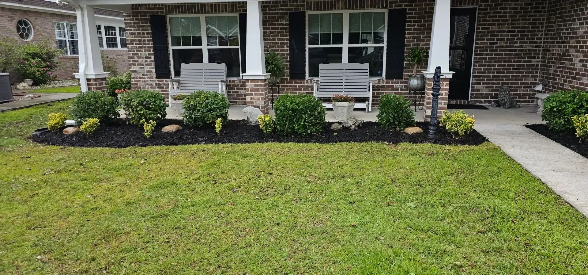 Green lawn with a brick house, a porch with benches and flowerbeds.