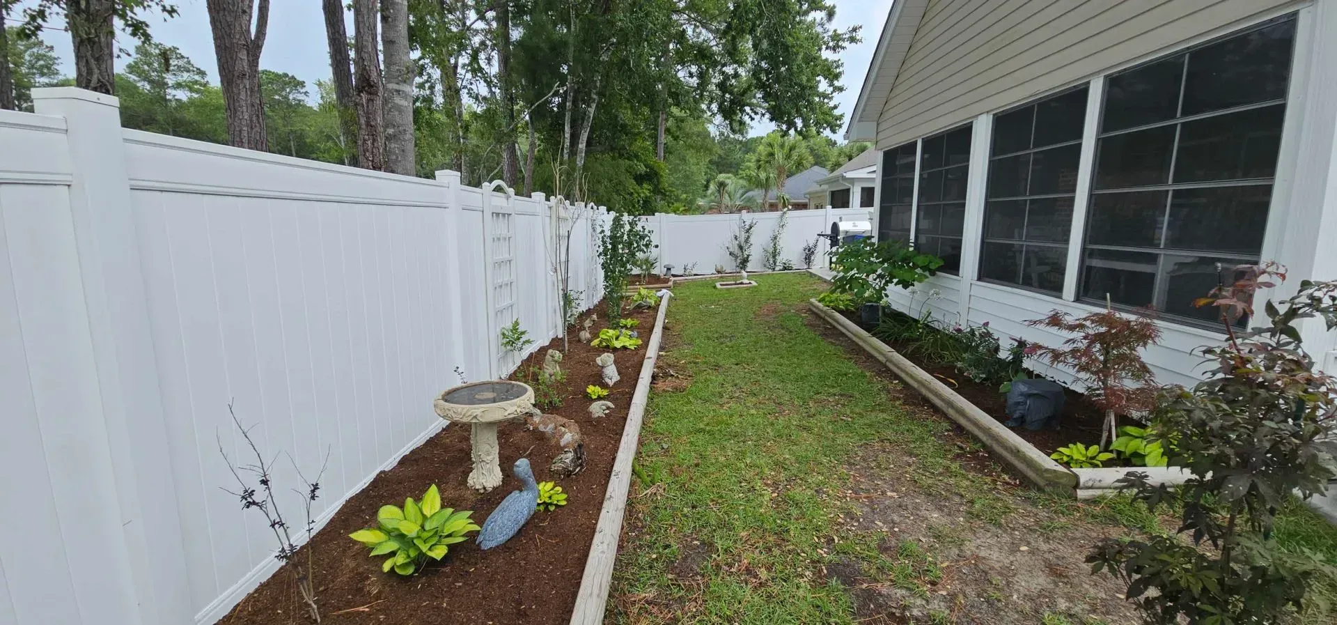 A backyard with a white fence, garden bed, bird bath, and grassy area next to a white house.