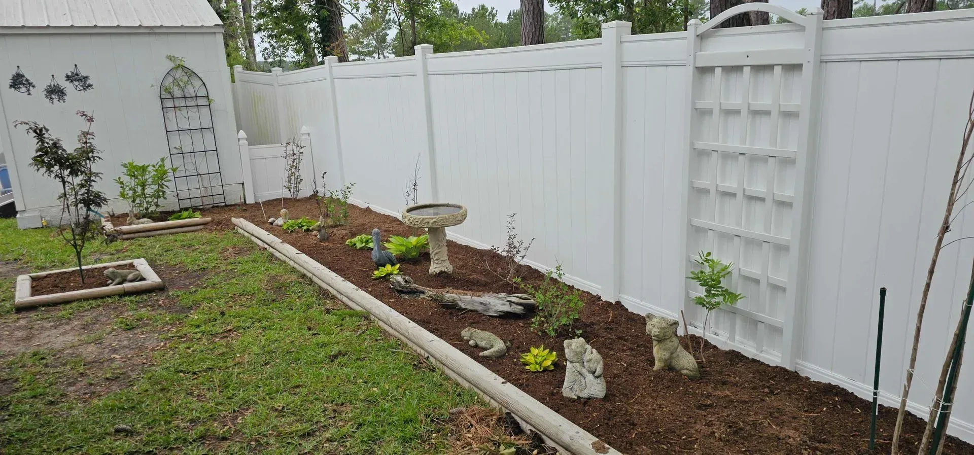 Backyard garden with white fence, birdbath, mulch, and small plants.