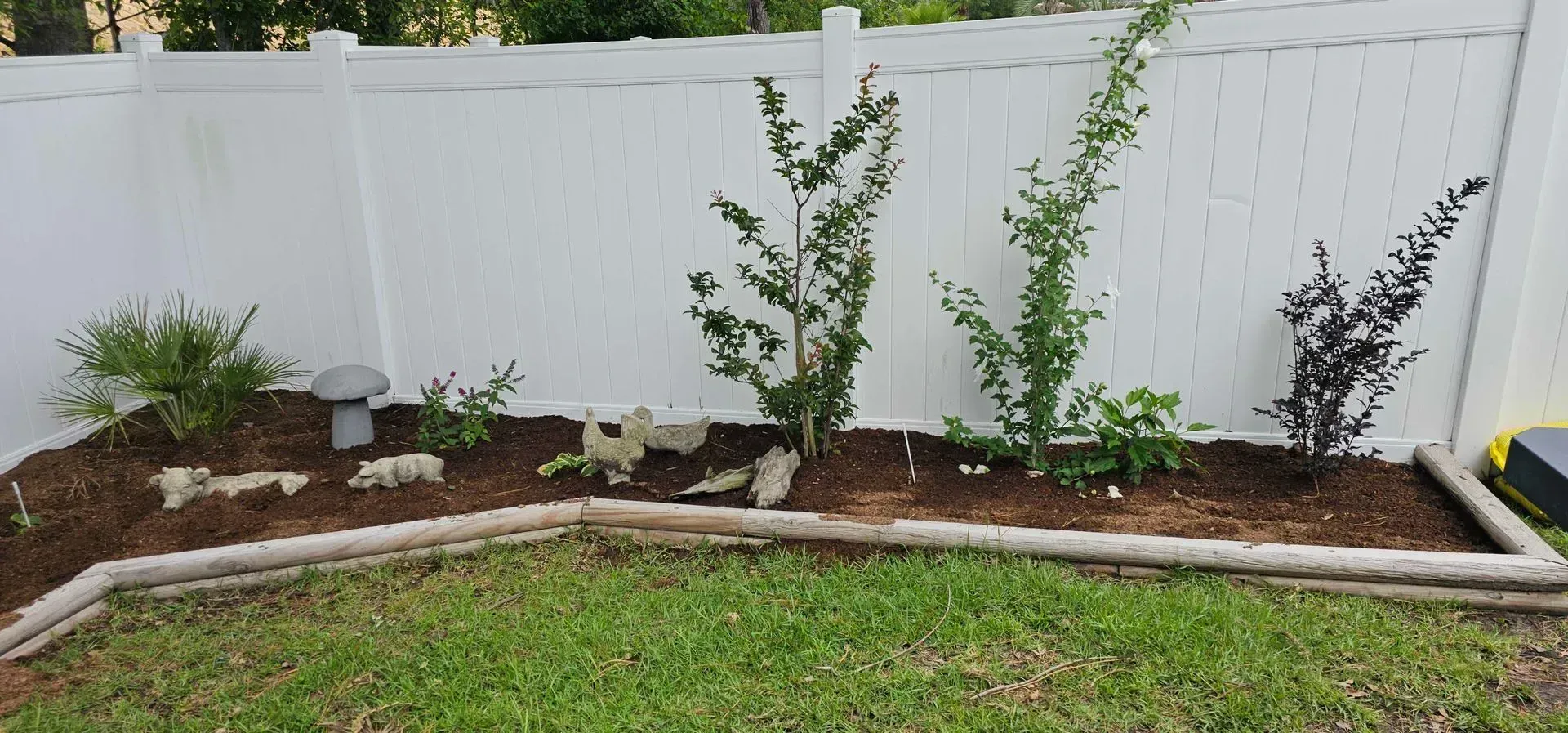 A backyard garden bed with small trees, rocks, and a mushroom statue in front of a white fence.