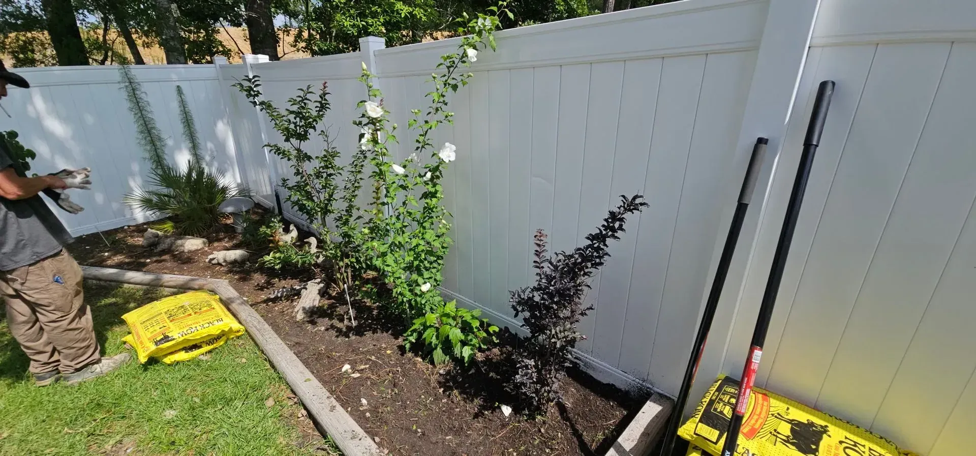 A person tending a garden along a white fence. Plants in a raised bed with tools nearby.