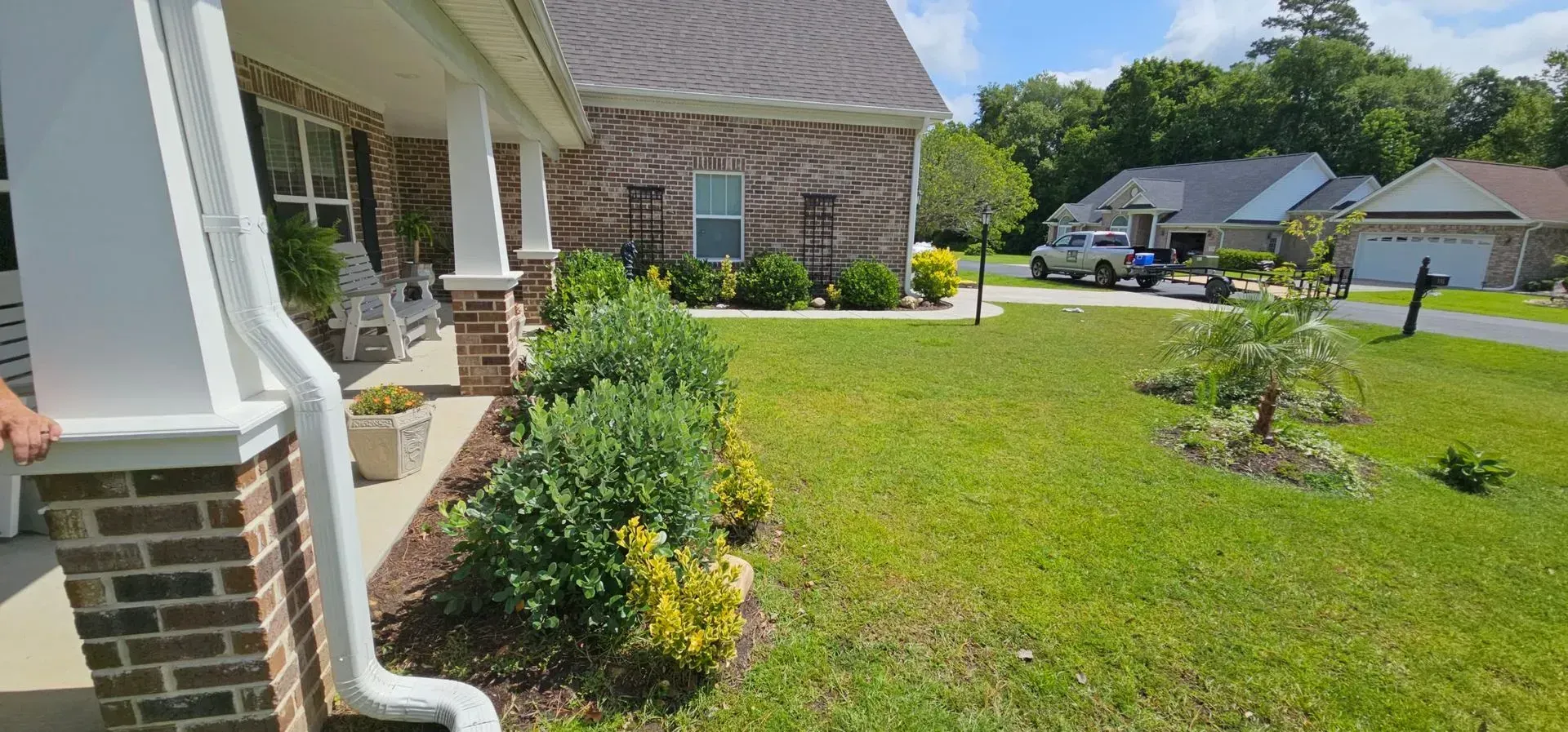 A brick house with a green lawn, bushes, and other houses in the background on a sunny day.