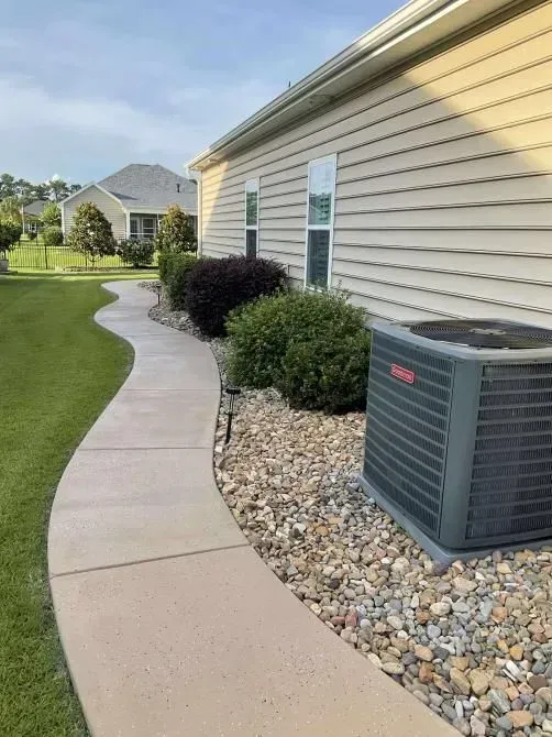 Concrete walkway curves past house with tan siding, bushes, and air conditioner.