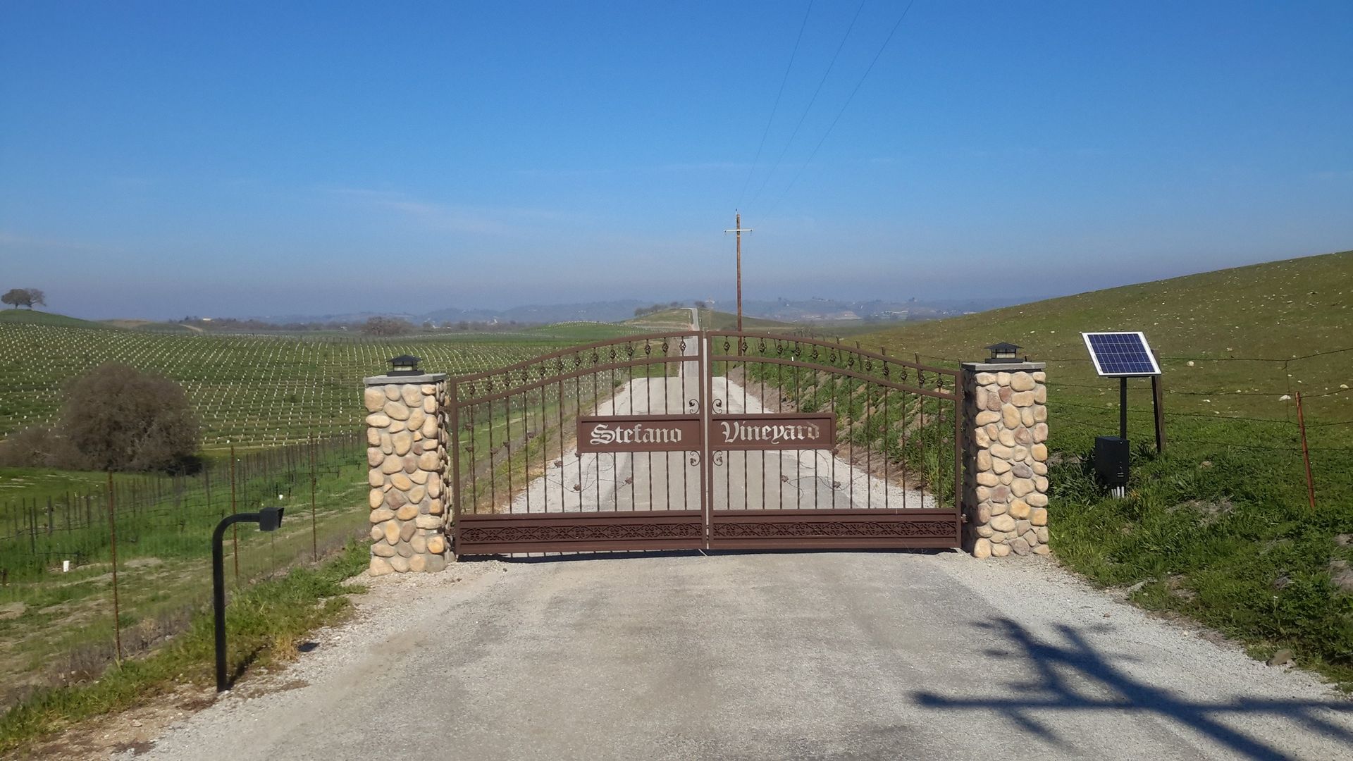A gated entrance to a vineyard, with a gravel road, solar panel, and fields in the background.