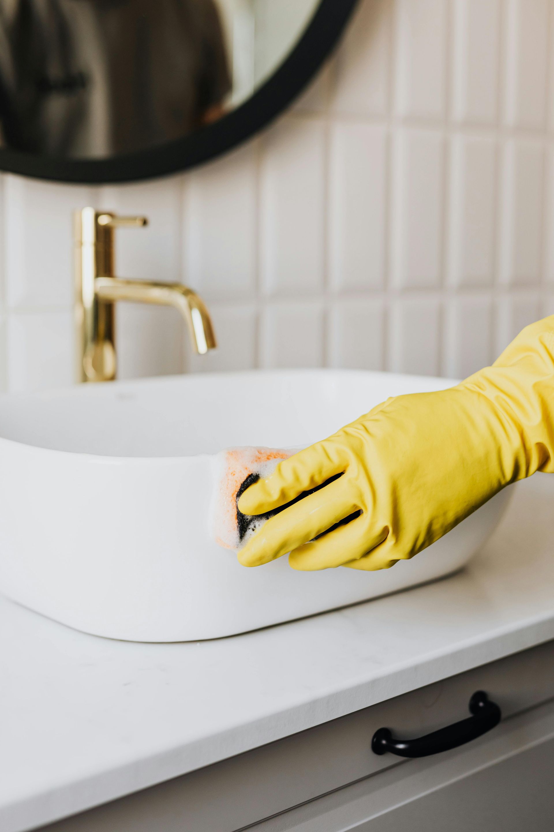A person wearing yellow gloves is cleaning a bathroom sink with a sponge.