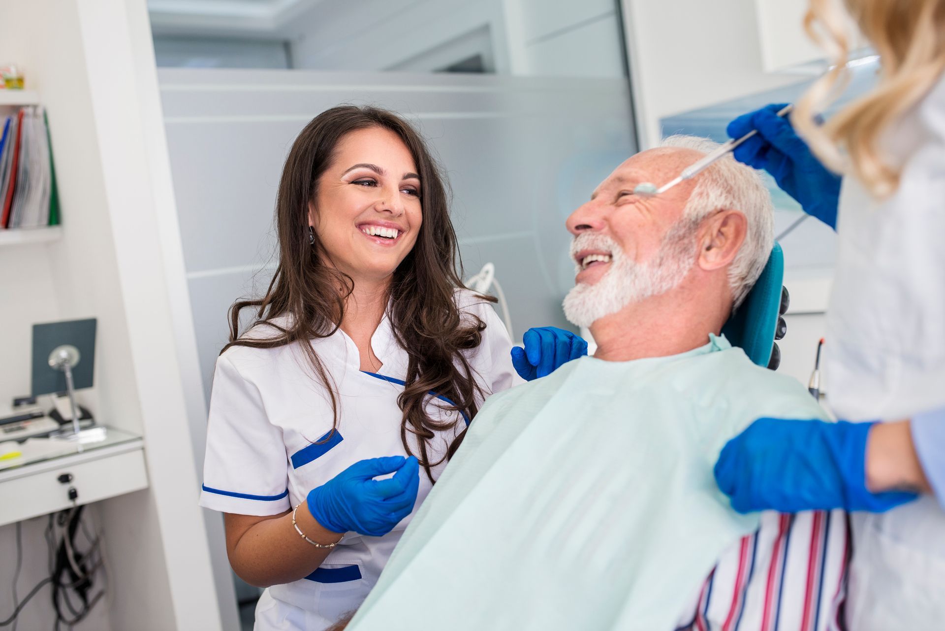 Male senior receives dental treatment from two female dentists as they all smile.