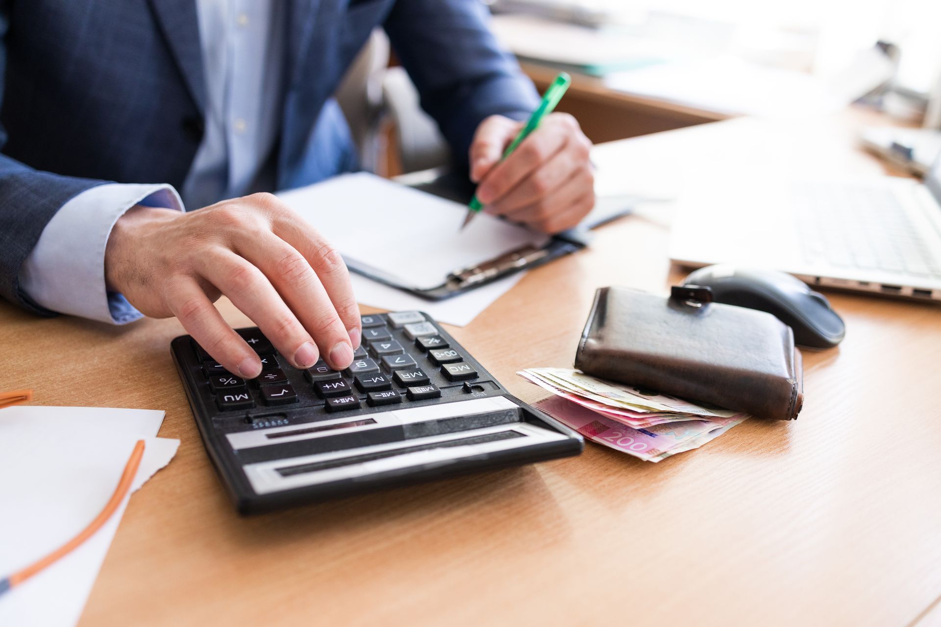 Man in suit uses calculator, writing on notepad, with wallet and cash on desk.