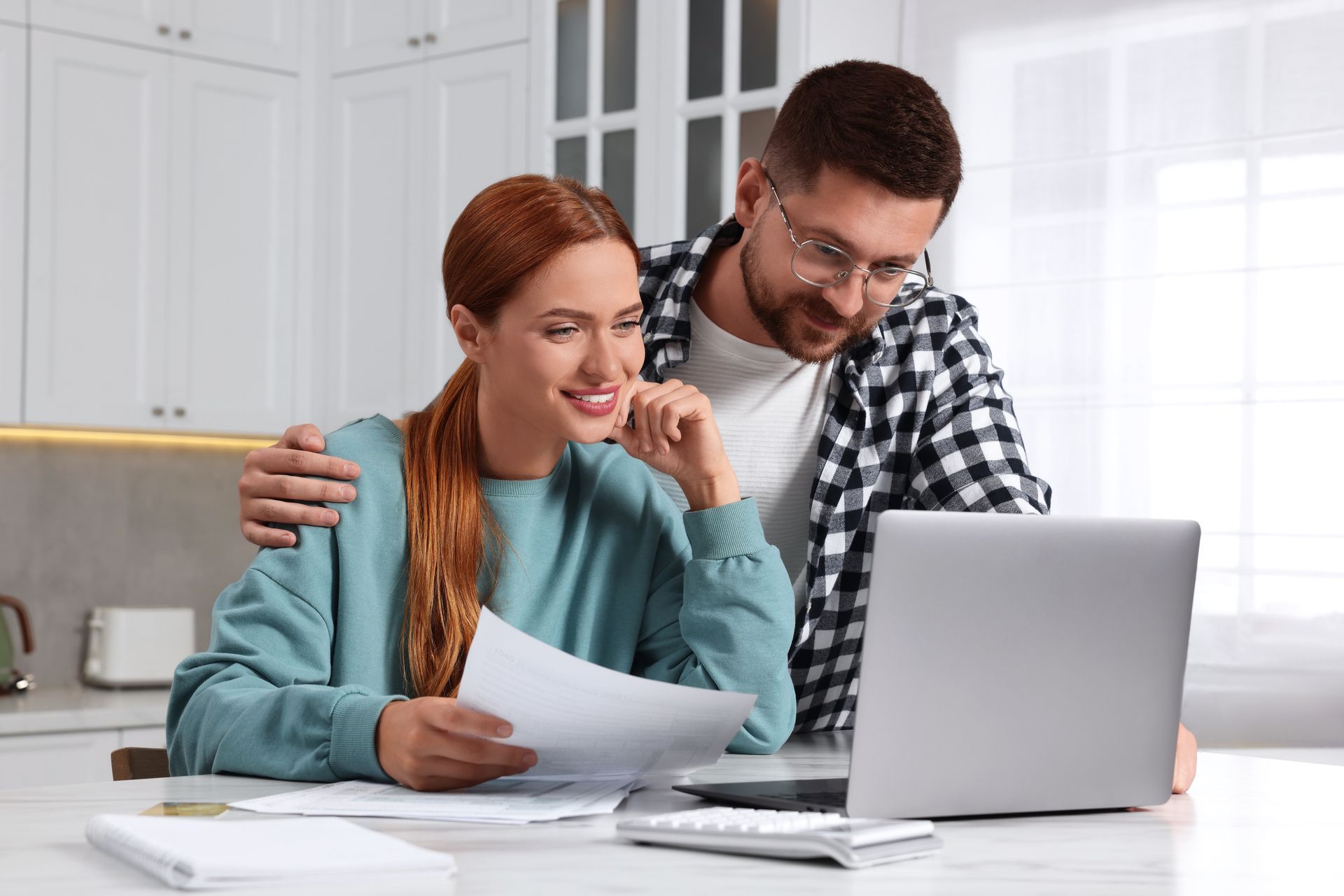 Couple reviews finances together at a laptop in their kitchen. Woman holds papers, both smile.