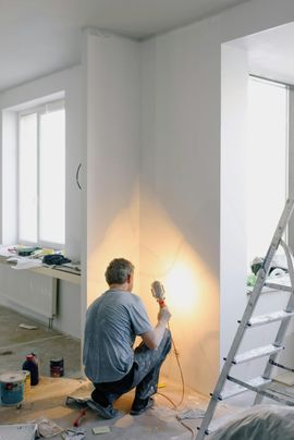 Painter kneeling by a wall, applying paint with a roller in a bright room beside a ladder