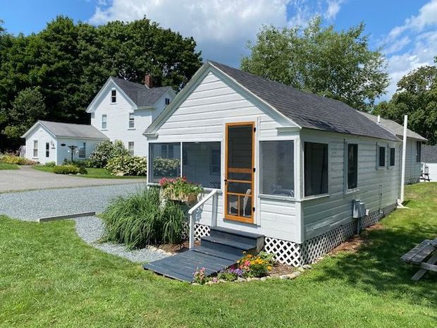 A small white house with a screened in porch is sitting in the middle of a lush green field.