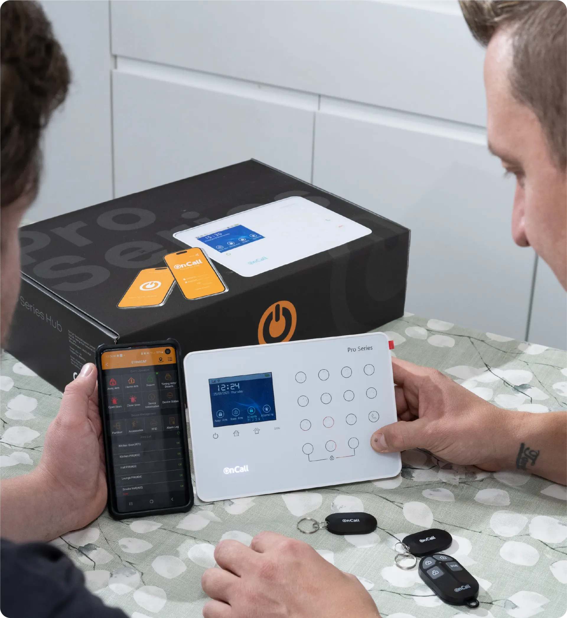 Two people interacting with a security system. One holds a phone with an app, the other a control panel.