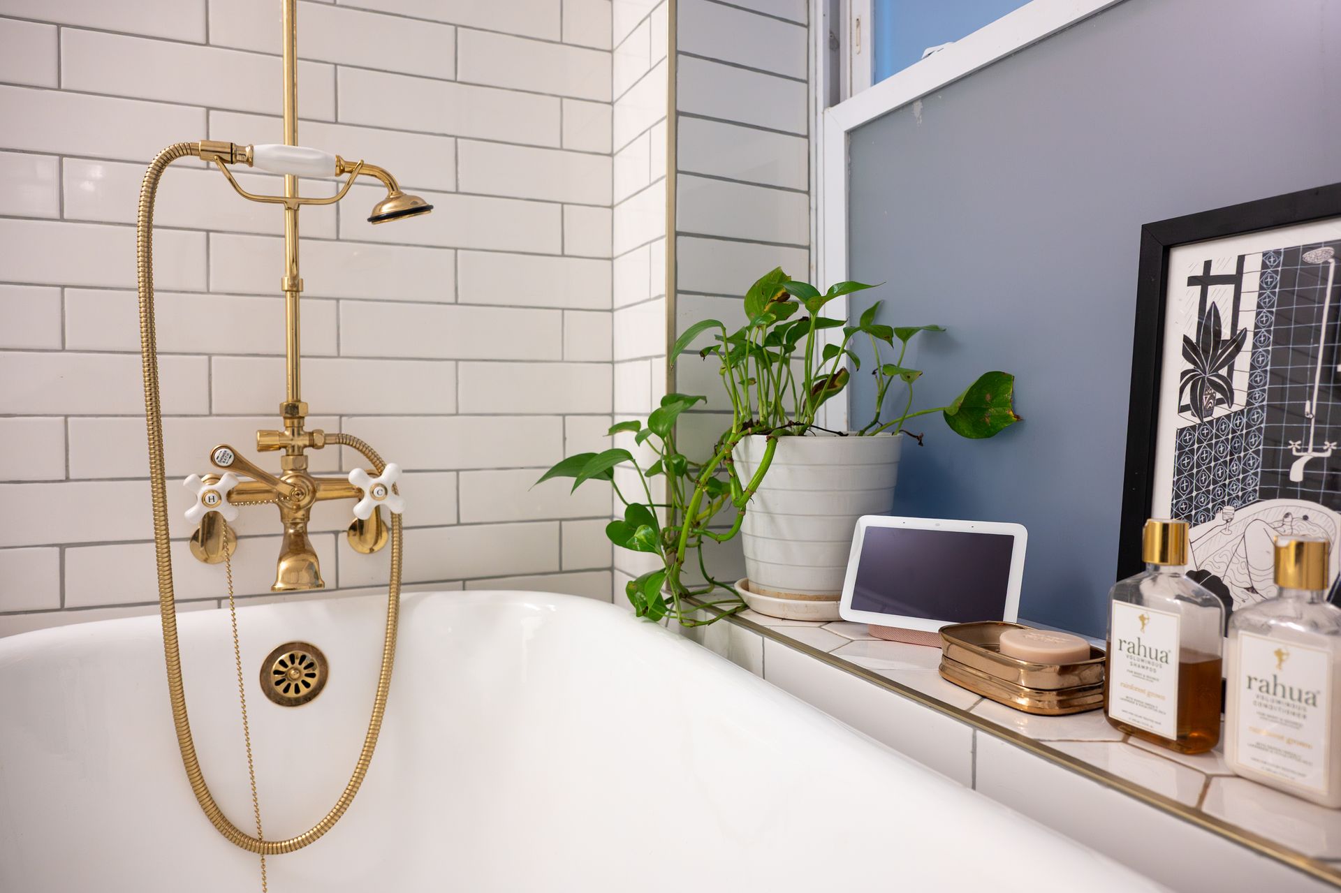 White bathtub with gold fixtures, surrounded by white subway tiles. Shelf with plant, framed print, and toiletries.