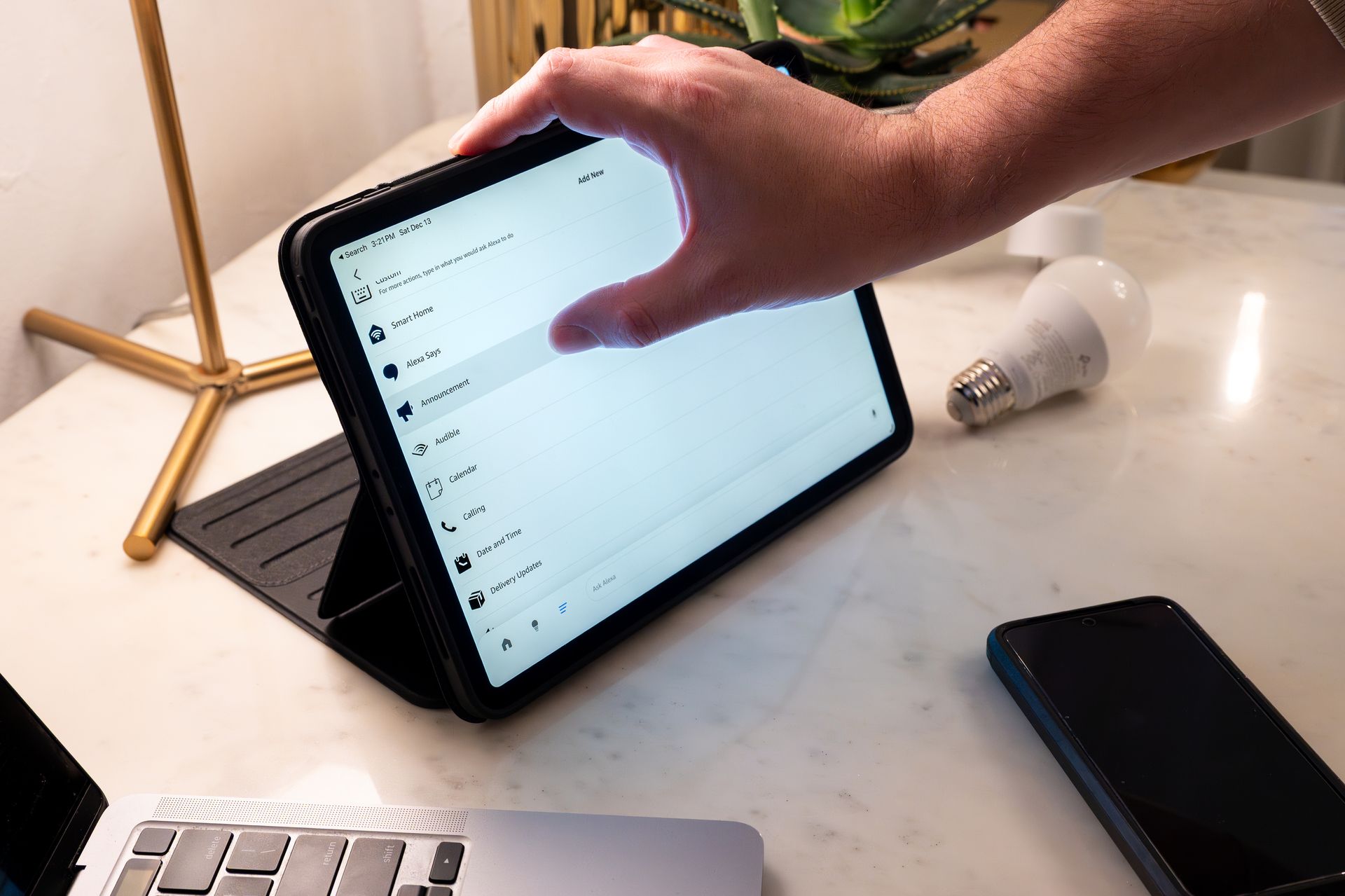 A hand touching a tablet screen on a stand. A lightbulb and phone are also on the table.