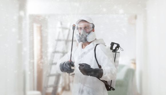 Pest control worker in a white suit, respirator, spraying inside a room.
