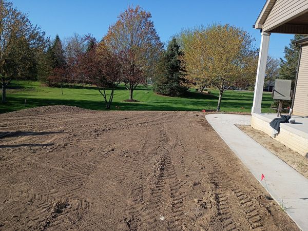 A bare dirt patch next to a concrete sidewalk near a house and a lawn with trees.