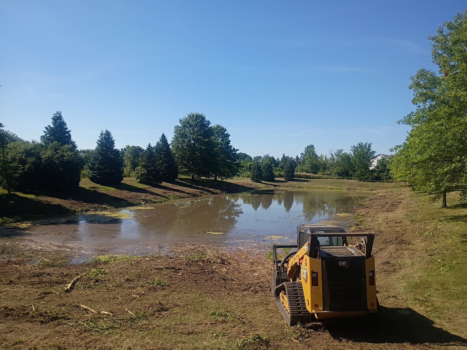 A yellow skid steer clears debris from the edge of a pond under a blue sky.