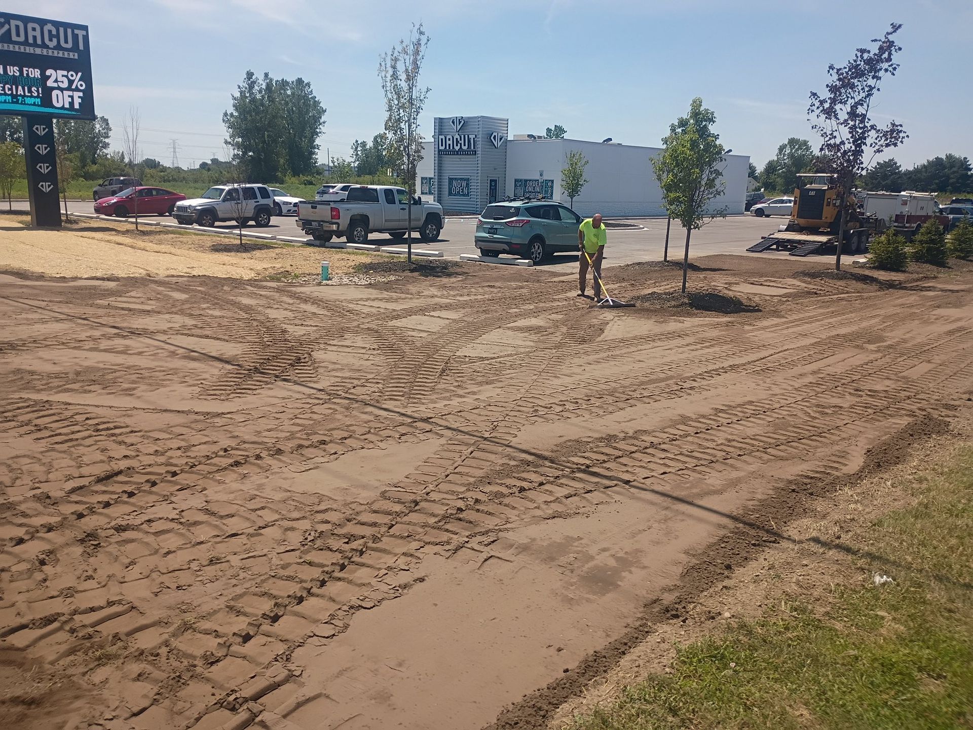 Construction site with dirt and tire tracks, worker in yellow vest, buildings and vehicles in background.