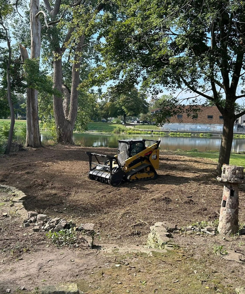 Yellow skid steer on mulch, near a pond and trees.
