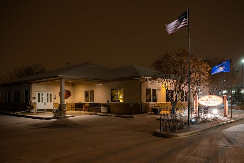 A beige, single-story funeral home at night with an American and blue flag on poles and a lit sign out front.