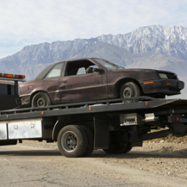 A dark-colored, aged coupe sedan loaded onto the flatbed of a tow truck, with mountains in the background.