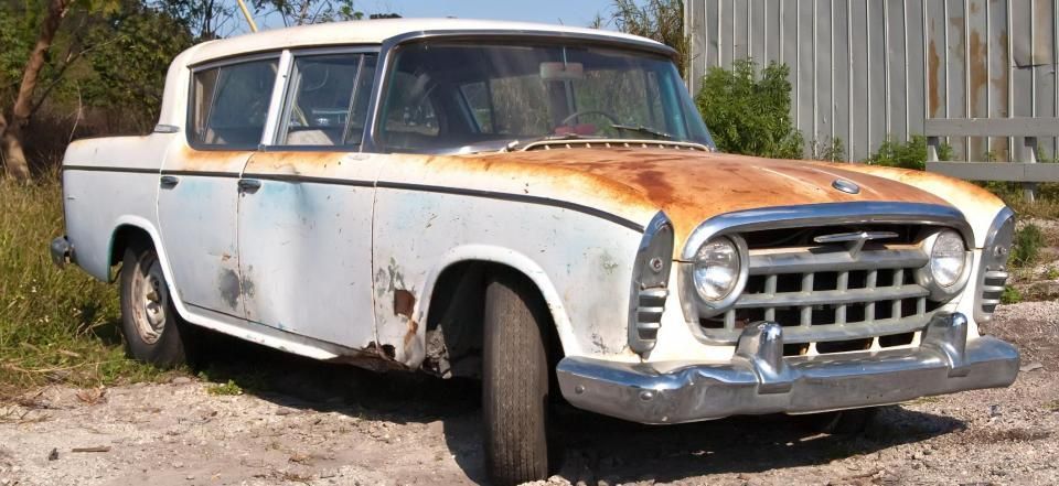 A weathered, white, rusted classic 1950s sedan parked on dirt in an outdoor setting.