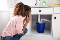 A woman is kneeling under a sink talking on a cell phone.