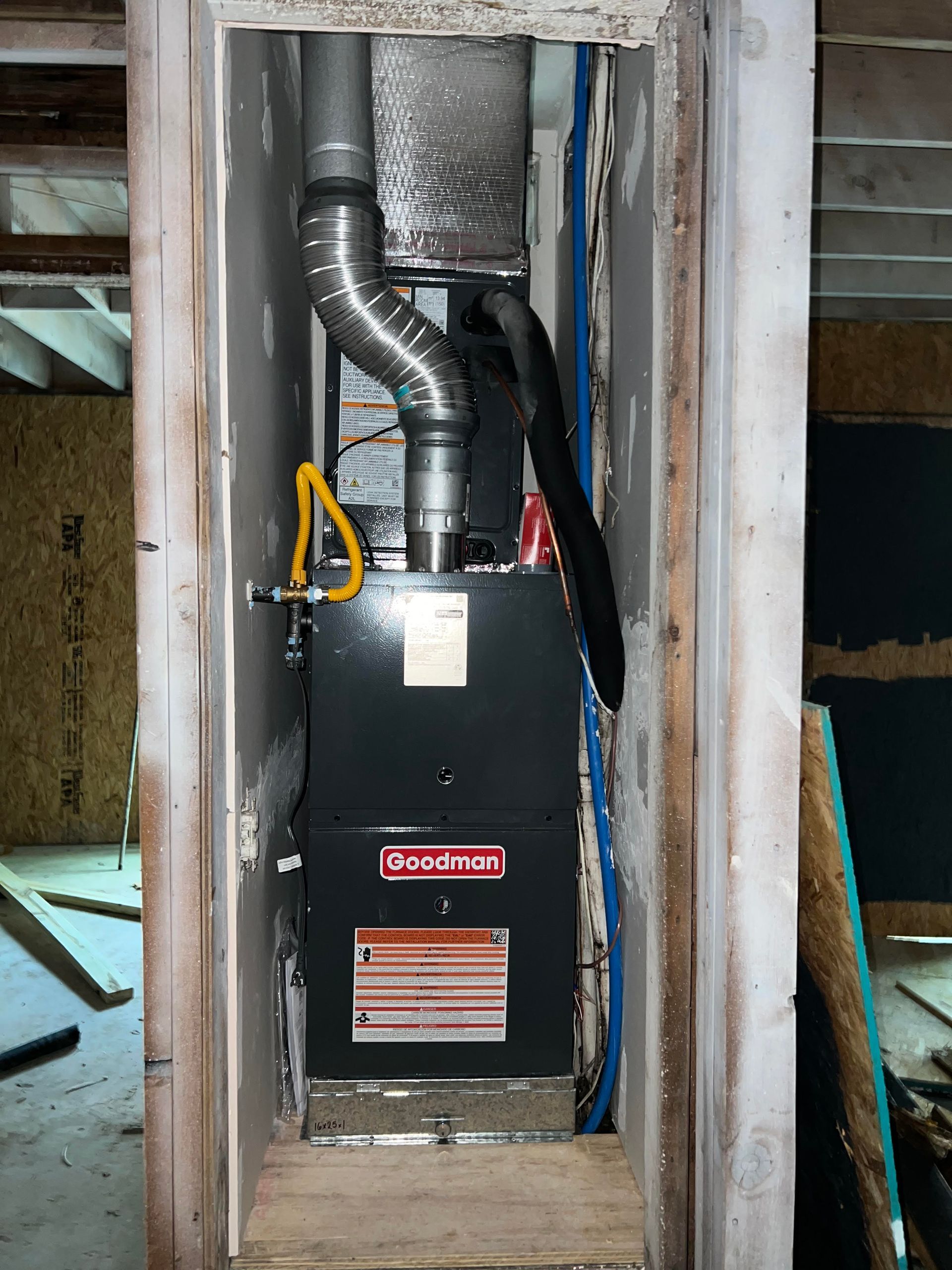 A Goodman furnace installed in a framed, unfinished wall cavity; silver ductwork, black unit with warning label.