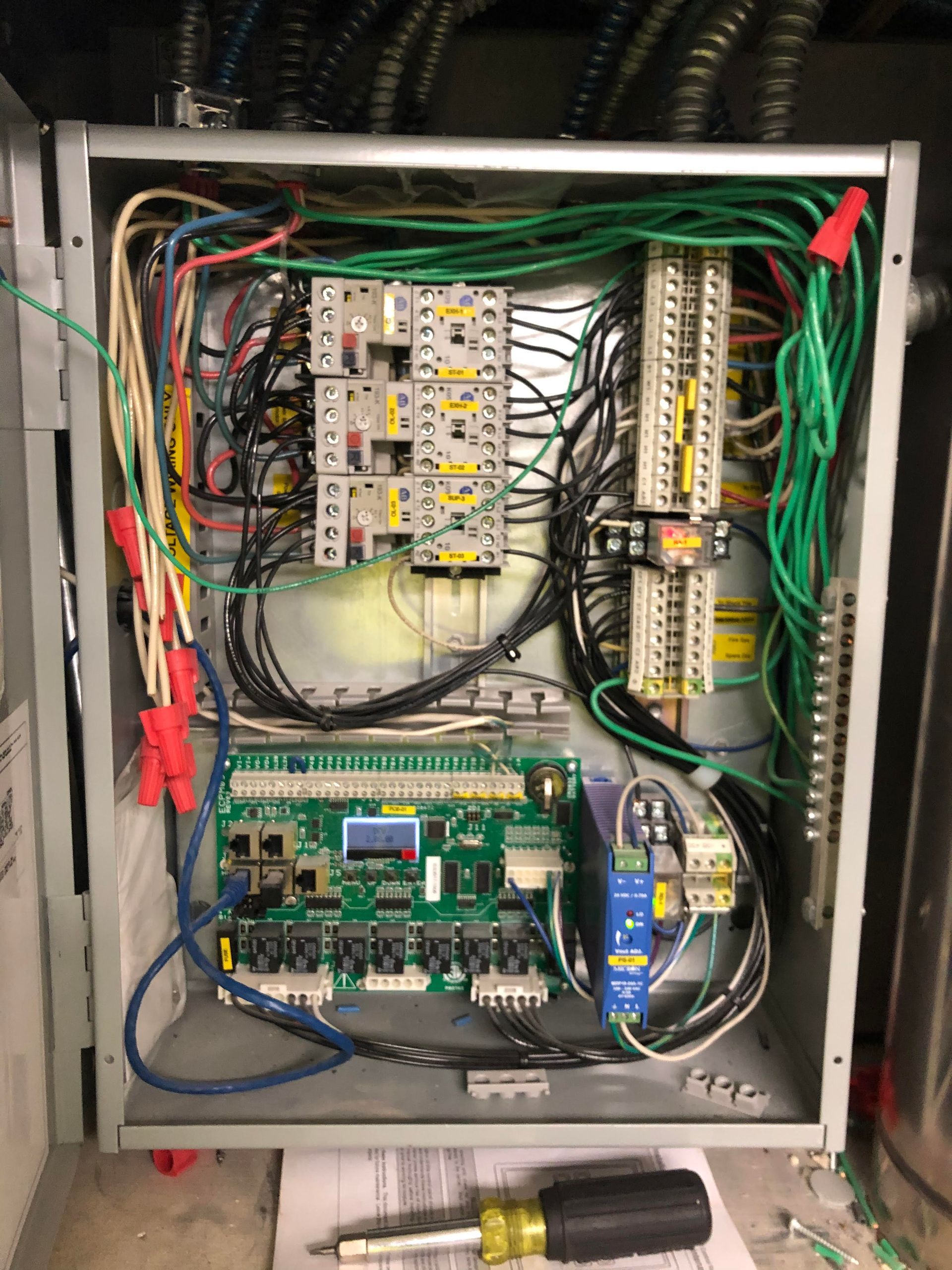 Open electrical panel with wires, circuit boards, and terminal blocks. A screwdriver rests below.