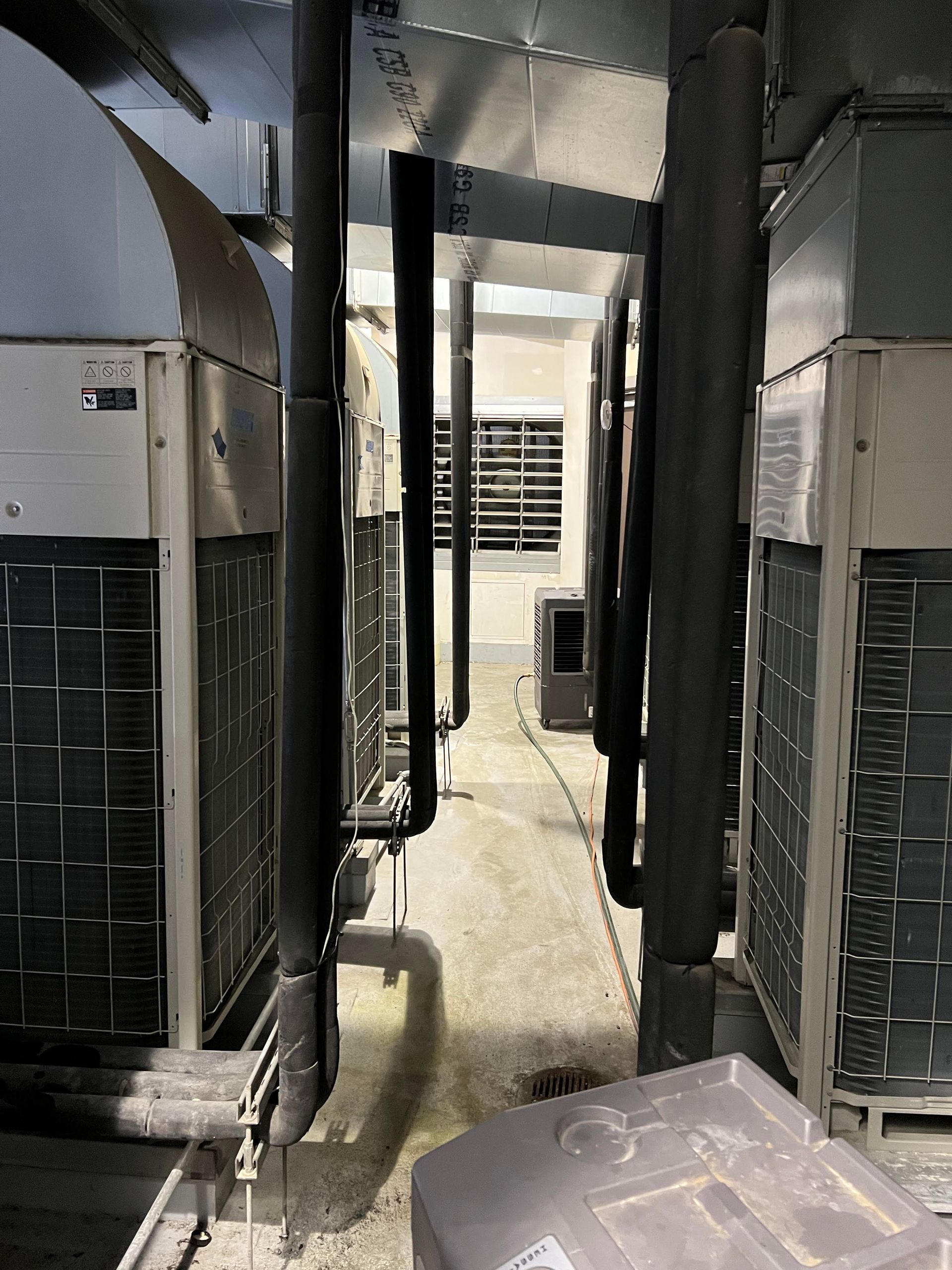 HVAC units and insulated pipes line a narrow concrete corridor, leading toward a distant, open doorway.