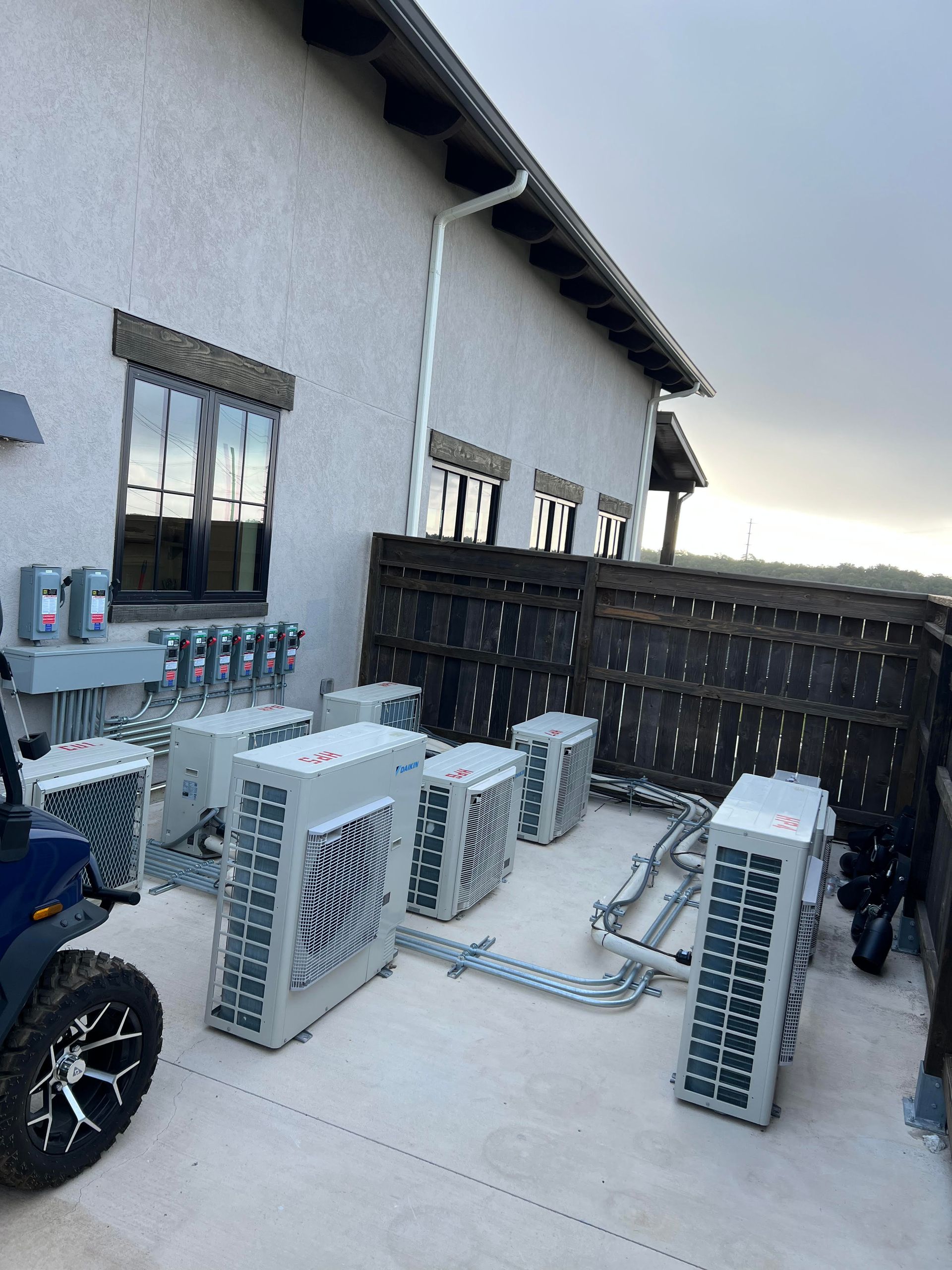 Outdoor air conditioning units near a building's exterior wall, beneath a roof and windows, connected by metal pipes.